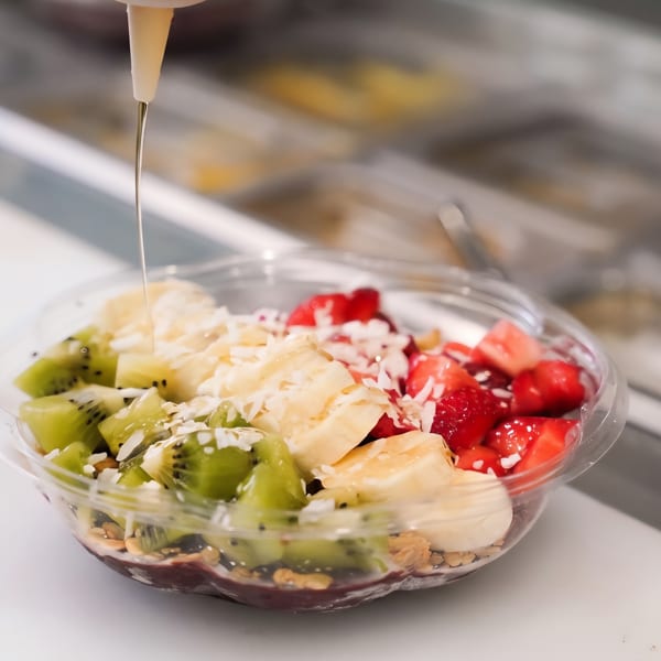 Fruit bowl topped with kiwi, strawberries, banana, shredded coconut, and syrup being poured over it.