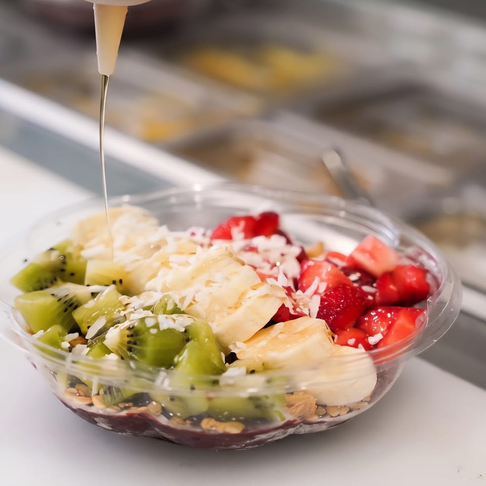 Fruit bowl topped with kiwi, strawberries, banana, shredded coconut, and syrup being poured over it.